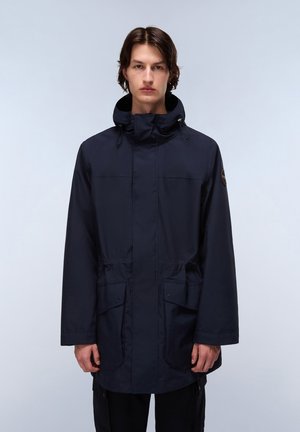 Jonge man in een donkerblauwe parka met capuchon en voorzakken, staande tegen een effen lichtgrijze achtergrond, recht vooruit kijkend.