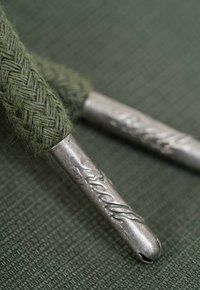 Cuerda trenzada verde con extremos metálicos, que presenta un logo grabado. El fondo es una tela de color verde oscuro con textura.