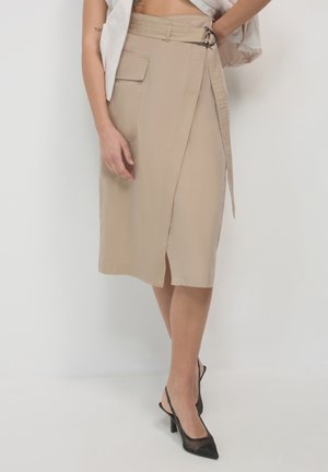 Donna che indossa una gonna avvolgente beige con tasca laterale e cintura, abbinata a scarpe con tacco alto a rete nere con cinturino alla caviglia, in piedi contro un muro bianco.