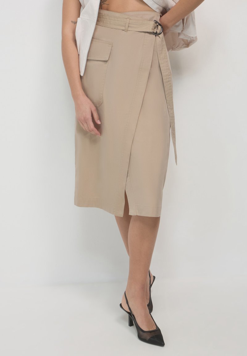 Femme portant une jupe enveloppante beige avec poche latérale et ceinture, associée à des chaussures à talons aiguille noires en mesh, se tenant contre un mur blanc.