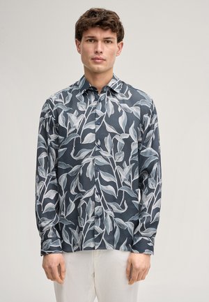 Junger Mann mit kurzen braunen Haaren, trägt ein graues Hemd mit weißen Blattmustern und weiße Hosen, steht vor schlichter Hintergrundkulisse.