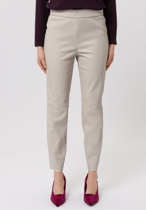 Woman wearing beige fitted trousers and burgundy pointed high heels, standing against a plain light background.