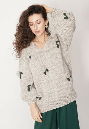 Gruby beżowy sweter z dekoltem w kształcie litery V, dekoracyjnymi aplikacjami w kształcie róż oraz zielonymi liśćmi na oversize'owych rękawach. Żebrowe wykończenie na dole i przy mankietach.