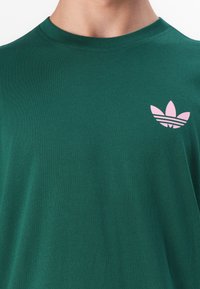 Zaļš kokvilnas T-krekls ar apaļu kaklu un rozā Adidas trīszaru logotipu kreisajā krūtī. Gluda tekstūra un regulāra piegriezuma.