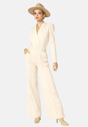 Femme portant une combinaison pantalon ample couleur crème, un chapeau beige, des boucles d'oreilles créoles dorées et des bottes à talons beiges, debout avec une main dans la poche.