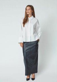 Chemise blanche à boutons avec des poignets larges, associée à une longue jupe en cuir noir avec des coutures visibles, combinée avec des talons hauts noirs.