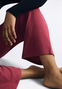 Rode geribbelde wijde broek met een cropped lengte en een klein logo detail aan de zoom. Een blote voet is zichtbaar op de voorgrond.