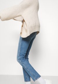 Ljust beige stickad tröja med avslappnad passform och sidoknappar, kombinerat med mörkblå jeans och vita slip-on skor. En enkel, avslappnad outfit.