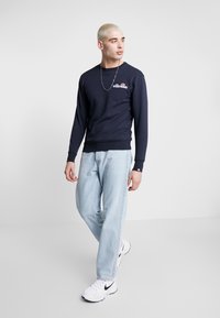 Sudadera de manga larga en color azul marino con cuello redondo y detalle del logotipo en el pecho. Combinada con jeans de color azul claro y zapatillas blancas.