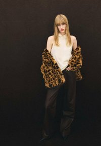 Ein Modell trägt ein cremfarbenes ärmelloses Top und schwarze weit geschnittene Hosen, umhüllt von einem braunen Leopard-Print-Faux-Fur-Mantel. Dunkler Hintergrund.