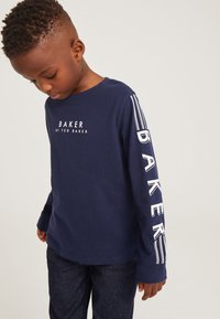 Navyblaues Langarm-T-Shirt mit weißem "BAKER"-Druck auf der Brust und dem Ärmel, verziert mit gestreiften Akzenten. Kombiniert mit dunklen Denim-Jeans.