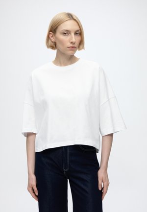 Kobieta z krótkimi blond włosami nosi oversize białą koszulkę i ciemne dżinsy, stojąc na tle jednolitego jasnego tła.