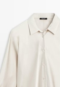 Camicia button-up beige chiaro con colletto a contrasto, realizzata in tessuto morbido. Presenta una fila di bottoni e una vestibilità comoda. Design semplice e classico.