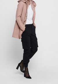 Ljusrosa huva jacka, vit t-shirt, svarta cargobyxor med fickor, och svarta stövlar med klack och genomskinliga detaljer.