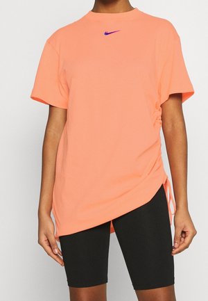 Kobieta ubrana w luźną, koralową koszulkę Nike z krótkimi rękawami i marszczeniem po bokach oraz czarne, dopasowane szorty sięgające do kolan.