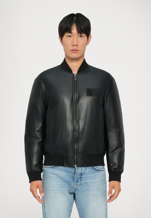 Čierna kožená bomber bunda s vysokým golierom, predným zipsom, rebrovanými manžetami a detailom náprsnej nášivky. Nosí sa cez svetlomodré džínsy.