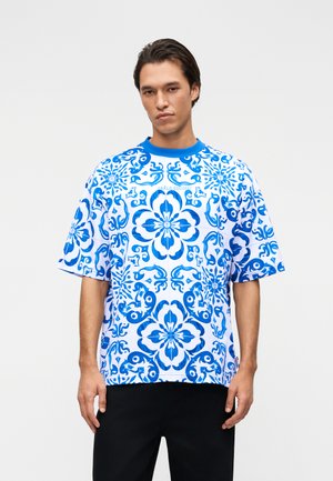 Mann trägt ein weißes kurzärmliges Hemd mit auffälligen blauen Blumen- und abstrakten geometrischen Mustern, steht vor einem einfarbigen hellen Hintergrund.