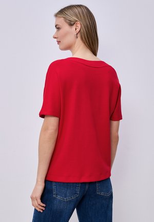 Vrouw met steil blond haar, gekleed in een rood topje met korte mouwen en een blauwe spijkerbroek, met de rug naar de camera en een neutrale achtergrond.