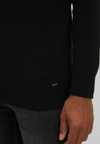 Schwarzer Strickpullover mit gerippten Bündchen und Saum, mit einem kleinen metallischen Markenetikett nahe dem unteren Saum. Nahaufnahme einer Hand, die den Ärmel hält.