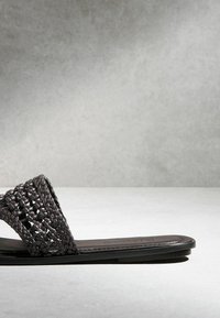 Sandale plate noire avec une large sangle en cuir tressé et une semelle fine sur une surface et un fond gris clair.