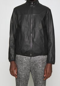 Svart skinnjacka med dragkedja, hög krage, elastiska muddar och två sidfickor. Bärs över texturerade grå byxor.