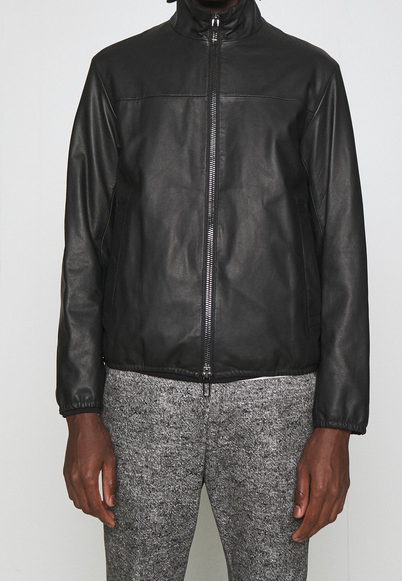 Svart skinnjacka med dragkedja, hög krage, elastiska muddar och två sidfickor. Bärs över texturerade grå byxor.