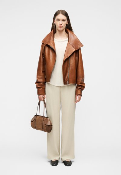 Γυναίκα στέκεται φορώντας υπερμεγέθη καφέ δερμάτινο σακάκι, κρεμ τοπ, φαρδιά μπεζ παντελόνια, μαύρα παπούτσια, κρατώντας καφέ τσάντα από το λουράκι στο δεξί χέρι.