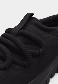 Κοντινό πλάνο μαύρου αθλητικού παπουτσιού με παχιές στρογγυλές κορδόνια και υφασμάτινη θηλιά στο επενδυμένο γλώσσα, που εμφανίζει ραφές στην επάνω επιφάνεια και στη σόλα.