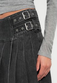 Fekete farmer szoknya redőkkel, széles derékpánttal, két ezüst csattal és szemekkel. Részben látható szürke hosszú ujjú felső.