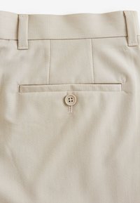 Pantalon beige avec une texture lisse, une poche arrière avec fermeture à bouton, et des plis subtils près de la ceinture.