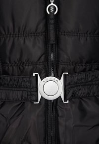 Chaleco negro tipo puffer con cremallera frontal y cintura elástica, acentuado con una hebilla circular de logo plateado. Textura suave y brillante.
