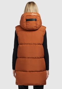 Persona bionda che indossa un gilet lungo senza maniche, imbottito, arancione ruggine con cappuccio, di spalle contro uno sfondo grigio chiaro uniforme.