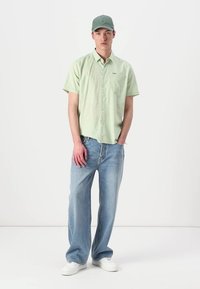 Camisa verde claro de mangas curtas com um bolso no peito, combinada com jeans azuis claros de corte largo e tênis brancos. Inclui um boné verde.