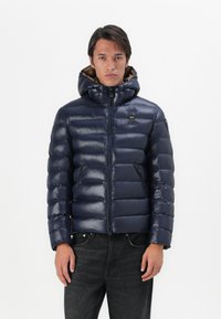 Marineblauwe gewatteerde jas met een glanzende afwerking, capuchon, ritssluiting en een horizontaal quiltpatroon. Bevat een logo op de borst.