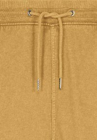 Cordon de serrage noué sur la taille d'un pantalon en tissu couleur moutarde, avec des embouts métalliques aux extrémités du cordon.