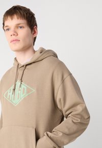 Beige kapucnis pulóver, pamutból készült, zöld, gyémánt alakú "HUGO" logóval, elülső zsebbel és kapucnis zsinórokkal. Sima textúra.