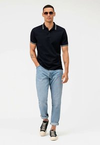 Polo blu navy con colletto a contrasto, jeans a risvolto azzurro chiaro e sneakers nere con lacci crema, indossati da una modella con occhiali da sole.