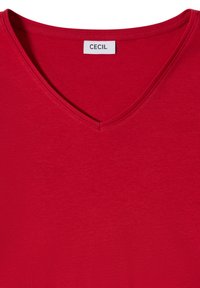 T-shirt rouge col en V avec une étiquette blanche à l'intérieur du col affichant le nom de la marque "CECIL" en lettres noires.