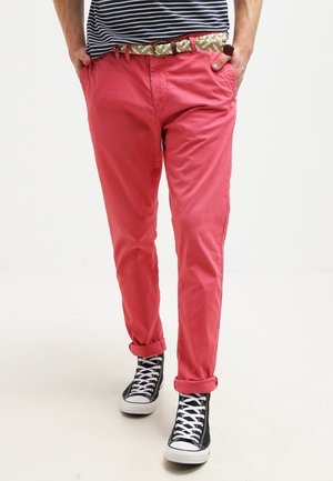 Osoba w różowo-koralowych spodniach podwiniętych do góry, czarnych wysokich trampkach z białymi sznurowadłami, koszuli w paski i plecionym pasku z klamrą w kształcie pierścienia.