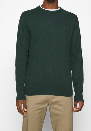 Personne portant un pull à manches longues vert foncé avec un petit logo sur la poitrine et un pantalon beige, debout devant un fond uni.