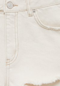 Valkoiset farkkushortsit, joissa on revitty helma, kultainen nappi, hienovarainen kudostehoste ja sivutaskut, joissa on vaaleita ompeluyksityiskohtia.