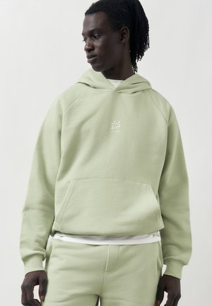 Młody czarnoskóry mężczyzna z dredami, ubrany w jasnokrótki zielony kaptur i pasujące spodnie dresowe, patrzący w lewo na jednolitym tle.