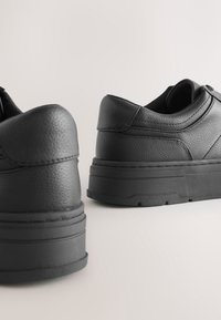 Paire de baskets en cuir noir avec semelles épaisses en caoutchouc, vues de dos sur un fond clair uni.