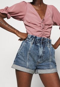 Blusa rosa de manga corta con cuello en V y detalle fruncido; pantalones cortos de mezclilla de cintura alta y dobladillo enrollado en azul con cierres de botón.