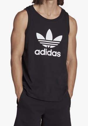 Personne portant un débardeur noir Adidas avec le logo tréfle blanc et du texte, mains dans les poches, cadré au-dessus des genoux.