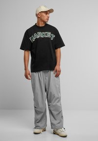 Svart oversized t-skjorte med "MARKET" i grønn skrift, kombinert med lysegrå, løstsittende bukser og kremfargede joggesko. Nøytralfarget caps.