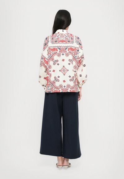 Witte blouse met een rood en marineblauw paisleypatroon, lange mouwen en een losse pasvorm, gecombineerd met marineblauwe wijde broek en hoge hakken.