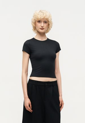 Γυναίκα με κοντά σγουρά ξανθά μαλλιά που φοράει εφαρμοστό μαύρο crop top με κοντά μανίκια και ψηλόμεσο μαύρο παντελόνι, στέκεται μπροστά σε απλή φόντο.