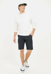 Witte langslavent shirt, marineblauwe cargoshorts met opgestikte zakken, en witte sneakers met marineblauwe accenten. Lichtblauwe pet met logo.
