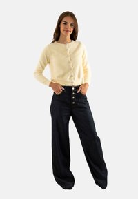 Cardigan de punto texturizado en color crema, con cuello redondo y botones abotonados, acompañado de jeans de pierna ancha de tiro alto en denim oscuro con botones dorados.